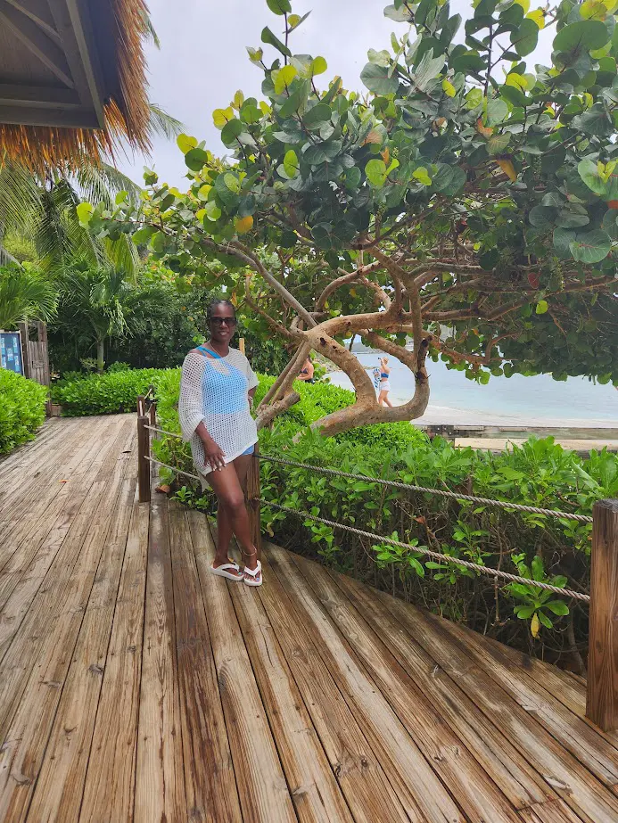 A woman in a white dress poses on a wooden deck by lush greenery and the ocean.