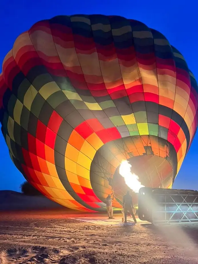 A colorful hot air balloon inflates at dusk with a bright flame.