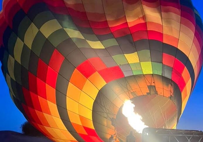 A colorful hot air balloon inflates at dusk with a bright flame.